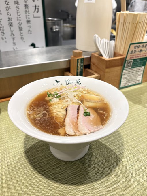 A bowl of Japanese ramen with chashu pork, menma, and scallions sits on a green placemat at a restaurant counter, with Japanese menu signs visible in the background.