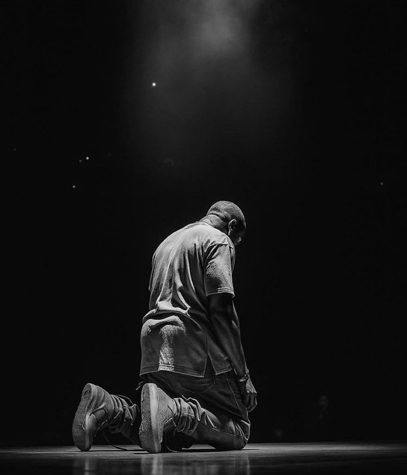 A dramatic black-and-white photograph of a man kneeling on a stage with his head bowed, illuminated by a single spotlight from above.
