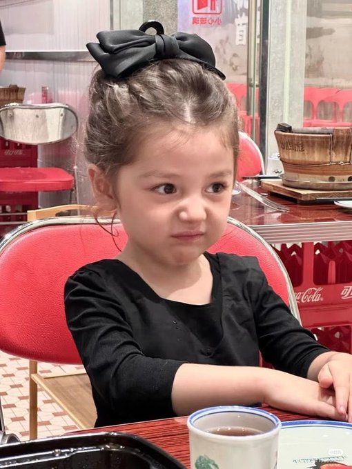 A young girl with a black bow in her hair sits at a restaurant table with a serious, determined expression, dressed elegantly in black. The image humorously accompanies the post about buying a memecoin, playing on the "little kid making big financial decisions" meme—suggesting the poster is jokingly as clueless (or as serious) as a child about to make a life-changing bet.