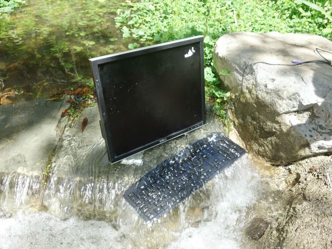 An abandoned computer monitor and keyboard placed in a shallow stream surrounded by green grass and stones, with water flowing around the electronics.