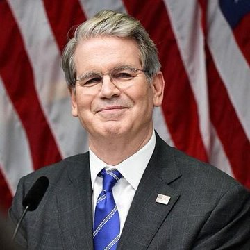 A man with gray hair and glasses, wearing a dark suit and a blue striped tie with a small American flag pin, smiles slightly in front of a U.S. flag backdrop. The image appears to relate to the Treasury's implementation of the GENIUS Act, possibly depicting Treasury Secretary Scott Bessent or a similar official overseeing the proposed stablecoin regulations.