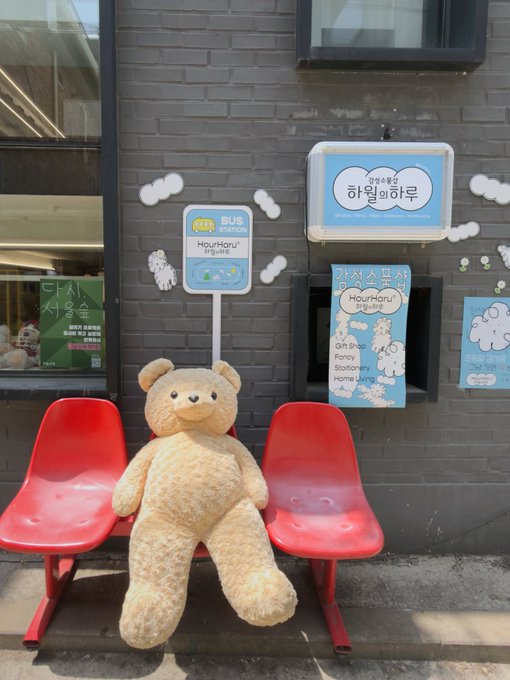 A large teddy bear sitting on a red bench between two other red chairs outside a shop with Korean signs including 'HaruHaru' and 'Gift Shop Fancy Stationery'.