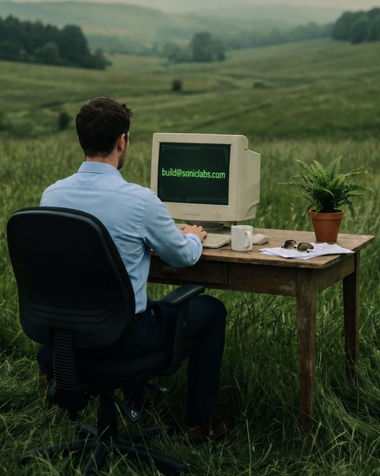 A man in a light blue shirt sits at a wooden desk with a retro CRT computer displaying 'build@soniclabs.com' in green text, set in a grassy green field under an overcast sky.