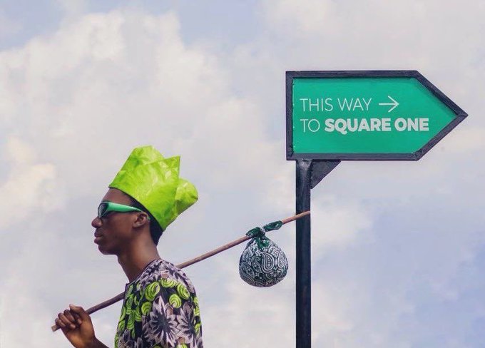 A person in green sunglasses, a green hat, and patterned clothing carries a pole with a small patterned bundle near a green arrow sign reading “THIS WAY → TO SQUARE ONE.”