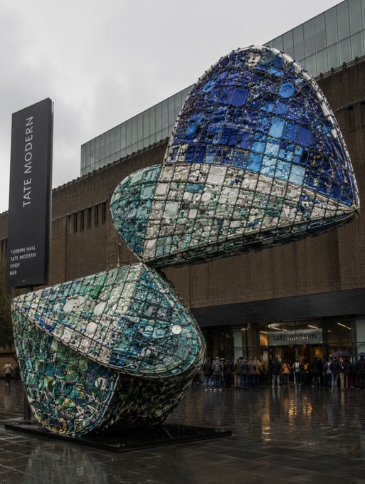 A large mosaic sculpture in shades of blue and green outside the Tate Modern museum in London, with the Tate Modern signage visible on the brick building behind it.