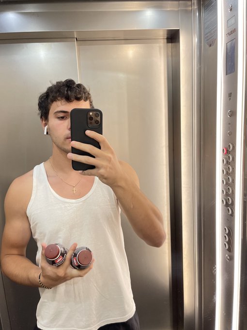 A man in a white tank top takes a mirror selfie in an elevator while holding two small jars.
