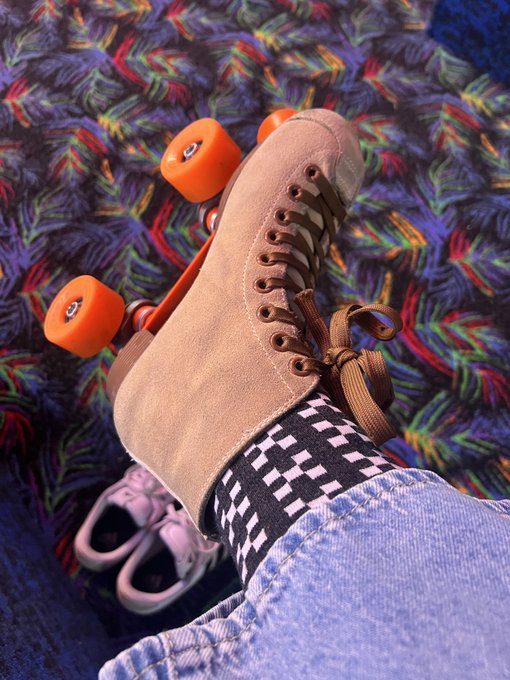 The image features a pair of blue jeans with white checkerboard patterned socks, both resting on top of roller skates. The person wearing these items appears to be posing for a photo or preparing to go skating. The caption "They say your OKX socks give you luck... and somehow i haven't fallen yet" suggests that the person might have experienced some luck in their life so far, possibly related to their choice of footwear with the checkerboard patterned socks.