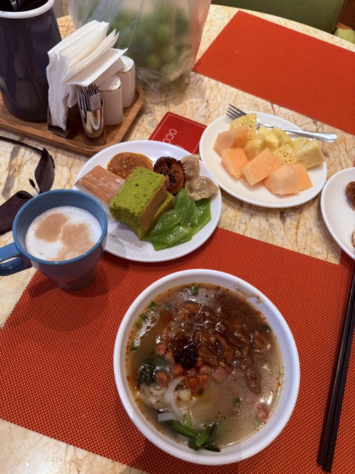 A breakfast spread on a table featuring a bowl of soup with meat and vegetables, a plate with a green cake, meat slices, and lettuce, a plate of assorted fruit, and a blue mug of coffee.