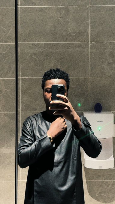 A man in a black leather-like shirt takes a mirror selfie with a smartphone in a public restroom.