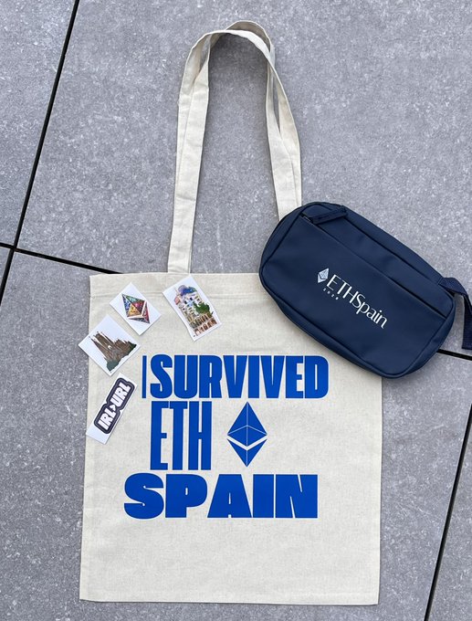A cream-colored tote bag reading 'I SURVIVED ETH [Ethereum logo] SPAIN' sits on a tiled surface alongside a navy 'ETH Spain' fanny pack and several stickers including 'LOCK ✕ PUNK'.