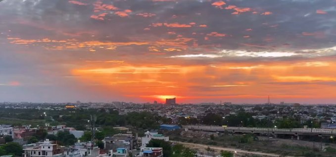 A scenic sunset view over a cityscape with pink and orange clouds illuminating the sky, featuring buildings, trees, and an overpass stretching across the horizon.