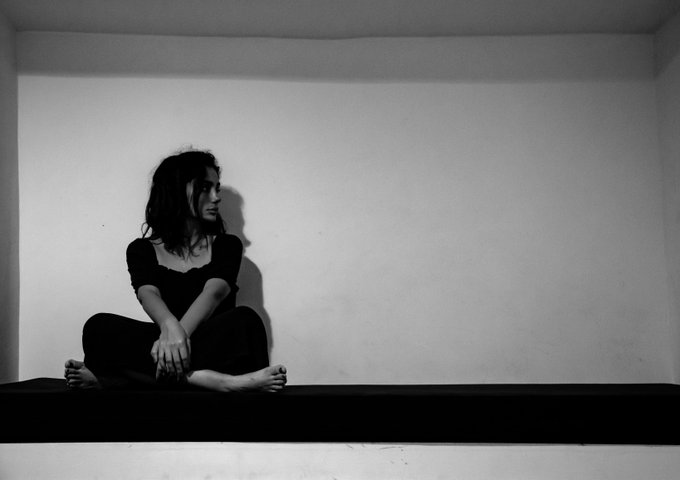 A black-and-white photograph of a woman sitting cross-legged in a sparse, white-walled room, looking off to the side with a contemplative expression.