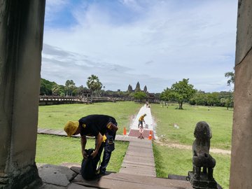 A view from a temple doorway at what appears to be Angkor Wat, showing a person in a yellow helmet adjusting their shoes on stone steps with the temple grounds and a central walkway visible in the distance.