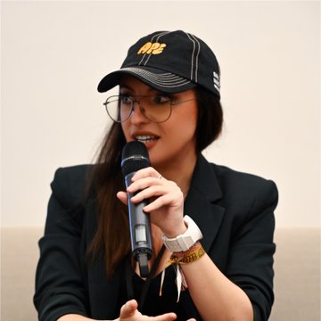 A young woman wearing glasses, a black Nike cap, and a black blazer speaks into a microphone while wearing a white Apple Watch.