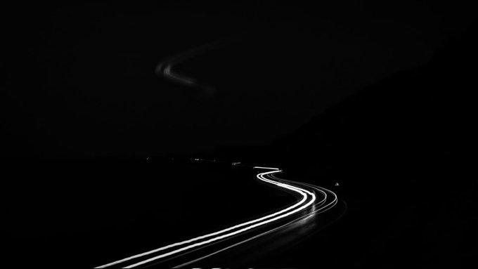 A long-exposure nighttime photograph capturing a winding road illuminated by streaks of white headlights, with a dark silhouette of a mountain and the faint glow of moonlight or a celestial body above.
