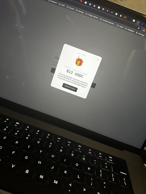 A laptop screen displays a Moonpay pop-up announcing a $12 USDC reward for claiming via the Stellar Tokenization Engine, directly matching the "$12 moon pay stimmy" referenced in the post. The keyboard is visible in the foreground, with the promotional gift box graphic on the screen reinforcing the crypto reward theme.