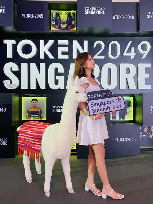 A woman in a short white dress stands beside a white alpaca draped in a colorful blanket, holding a 'TOKEN 2049 Singapore Summit' sign in front of a 'TOKEN 2049 SINGAPORE' backdrop.