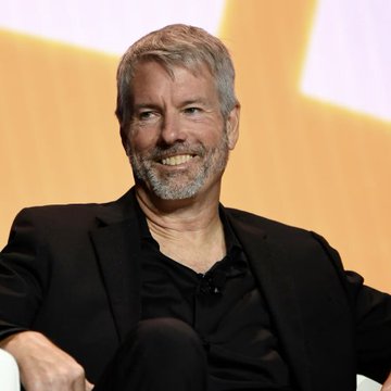A smiling man with a gray beard and short hair wearing a black suit jacket over a black shirt, seated at what appears to be a conference or panel event.