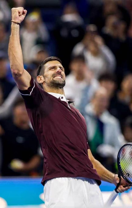 Tennis player Novak Djokovic in a maroon shirt celebrating with his fist raised and racket in hand, with a blurred crowd in the background.