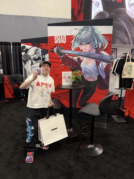 A man wearing a "Azuki" hat and shirt poses with an Azuki shopping bag in front of an Anime Expo booth featuring a large "Shio" character display. The image illustrates the post's caption, showing the Azuki TCG team member on-site at Anime Expo promoting their trading card game to new audiences.