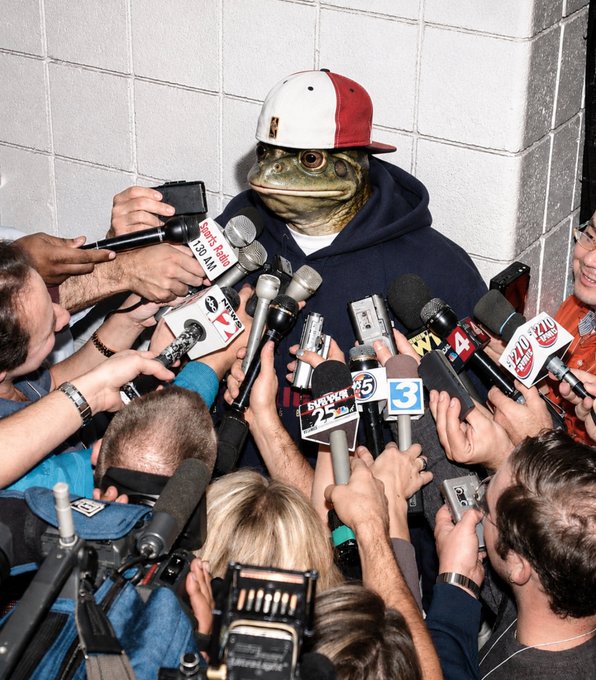A surreal, AI-edited or composited image showing a figure wearing a red and white baseball cap and a navy 'COLORADO' hoodie, but with a frog-like face, being interviewed by a swarm of reporters holding microphones from outlets like Sports 25, KUSA 9, 7, 3, and 4.