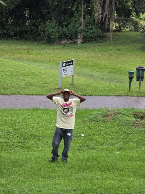 A young man in a cream t-shirt, jeans, and white cap stands on a grassy park area with his hands on his head, with a 'Hole #1' golf sign and ball washers visible in the background.