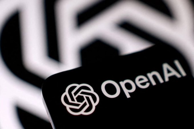 A close-up image of the OpenAI logo displayed on a black smartphone screen, with a blurred larger OpenAI logo in the background.