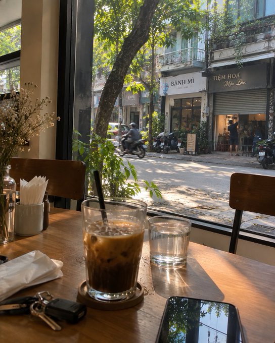 A view from inside a café looking out the window at a sunny street scene, showing a glass of iced coffee on a wooden table in the foreground, with a 'BÁNH MÌ' shop and 'TIỆM HOA' (flower shop) visible across the street in what appears to be Vietnam.