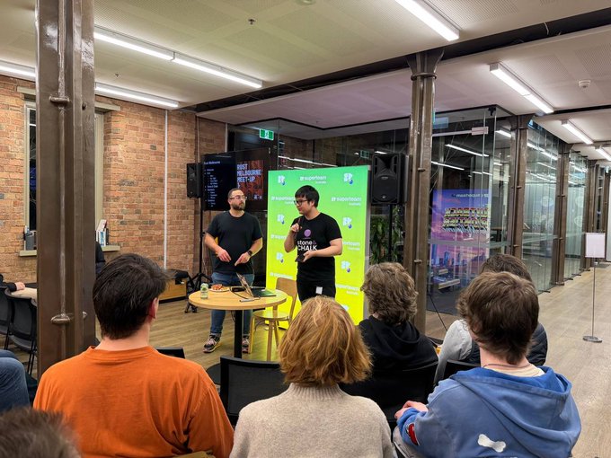 Two presenters stand on a small stage in a modern, industrial-style venue with exposed brick and metal pillars; one speaks into a microphone while the other looks on, as an audience watches from seated positions, with a bright green backdrop displaying 'Padel Network' branding.