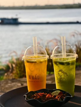 Two plastic cups with dome lids and straws, one containing an orange drink labeled 'X ĐẬU' and the other a green drink, sitting on a tray with a small dish of snacks, overlooking a body of water at dusk.