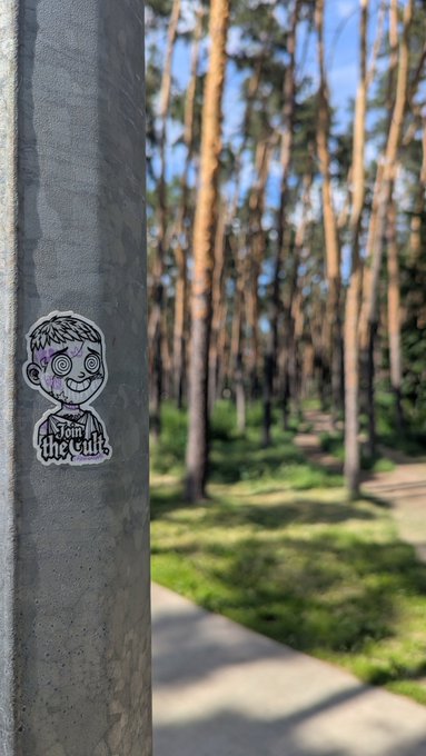A sticker of a cartoon boy with the text "Join the cult" is attached to a metal pole, set against a serene forest backdrop, fitting the weekend rest message shared by PytheniansNFT.
