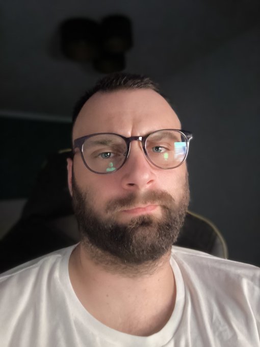 The image features a man wearing glasses, sitting down with his chin resting on his hand. He has a beard and is dressed in a white shirt. The background of the photo appears to be a dark room or a black background, which contrasts with the man's attire. There are no texts or other objects visible in the image.
