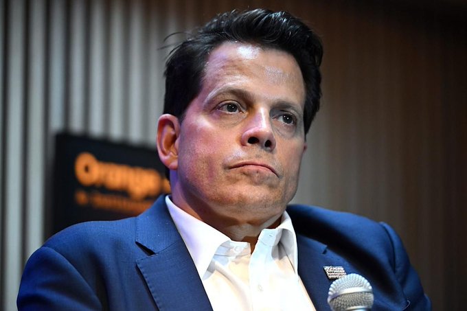 The image features a man wearing a suit, sitting at a table with his hands resting on his lap. He appears to be looking off into the distance or deep in thought. The background of the image shows an orange sign that reads "Orange's" along with some text about Bitcoin and its value.