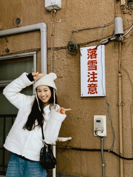 A woman wearing a white winter coat and beanie poses in front of a wall with a red and white sign with Japanese characters reading "落雷注意" (Beware of Lightning).