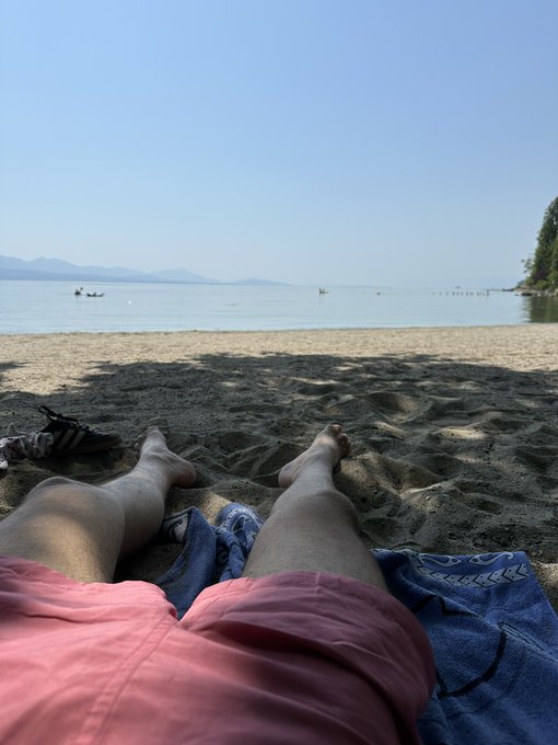 A first-person view shows legs stretched out on a sandy beach under a clear sky, with the calm water and distant mountains visible in the background.