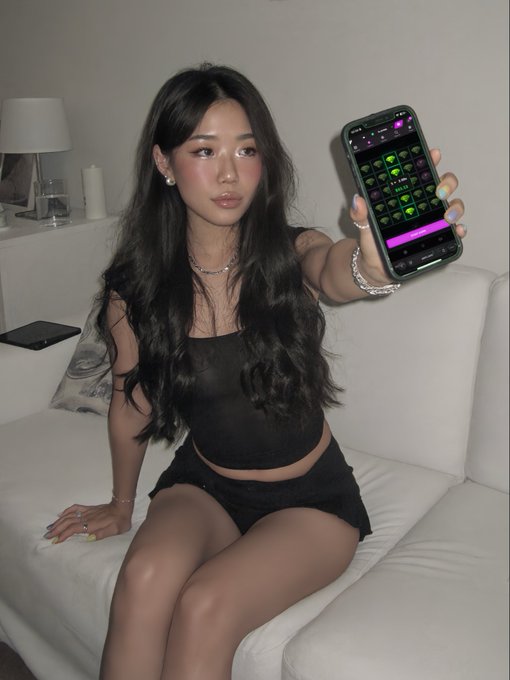 A young woman with long dark hair, wearing a black sheer top and skirt, sits on a white couch and holds up a smartphone displaying a heart-themed soundboard app interface.