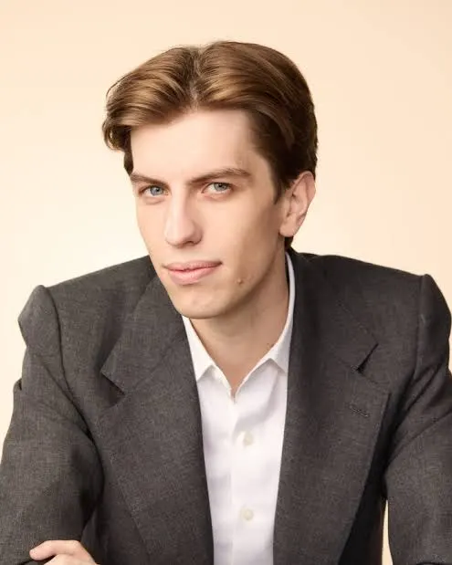 The image shows a young man with brown hair, wearing a gray suit jacket over a white dress shirt, posed in what appears to be a professional headshot against a light background. The image likely depicts Leopold Aschenbrenner, the founder of the AI-focused hedge fund Situational Awareness mentioned in the post, whose supposedly successful long-term portfolio collapsed due to 4x leverage in the final days of FTX.
