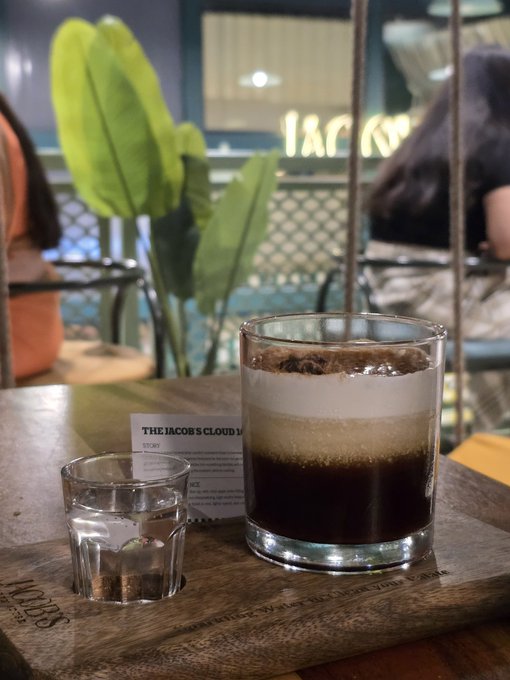 The image captures a coffee beverage, "The Jacobs Cloud Latte," served on a wooden tray with a glass of water, likely taken at a café to symbolize a luxurious or indulgent moment—fitting the post's theme of converting crypto USDT earnings into real-world purchases like a fancy coffee.