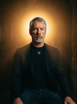 The image features a man sitting down with his arms crossed, wearing a black jacket and blue jeans. He has a serious expression as he looks directly at the camera. The background appears to be a dark room or an office setting, which contrasts with the bright light shining on him from above. There is also text in the image that reads "$MSTR The Monster has its upgrade. The skeptics continue to try and dismiss Saylor and the management team, yet continually fall short. Congratulations to shareholders who voted in favor of the $STRC amendment".