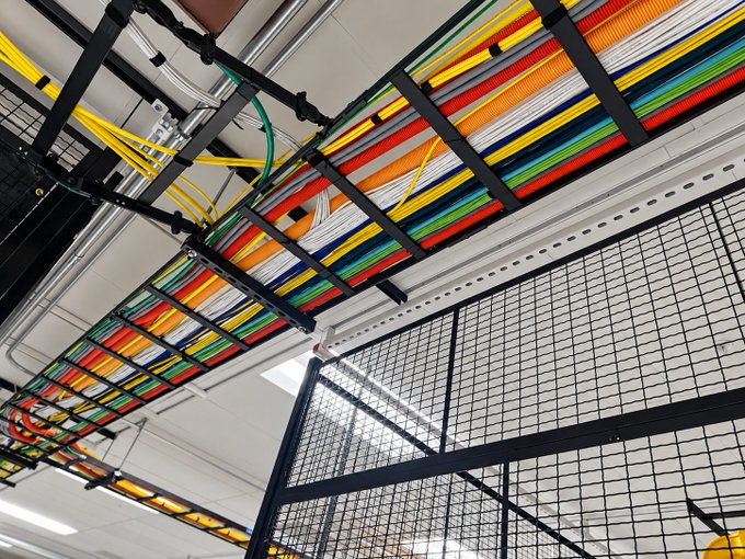 An industrial ceiling view showing an organized cable tray with multi-colored wires (yellow, orange, green, blue, red, white) running across it, with a black metal mesh fence below.