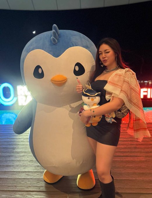 A woman wearing a black top and sheer shawl poses next to a large penguin mascot and holds a small penguin plush toy.