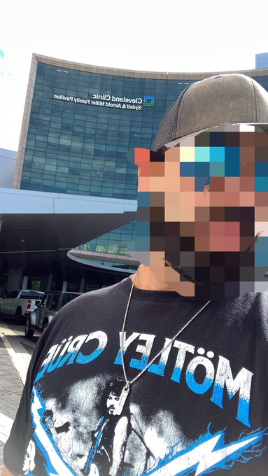 A man wearing a Motley Crue band shirt stands outside Cleveland Clinic Abu Dhabi, which advertises "Heart & Vascular Institute Pavilion," as he shares a moment before his 70-year-old mother's emergency open heart surgery, calling his mom "the true Bitcoin" in this deeply personal update.