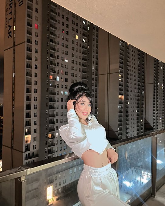 A woman in a white long-sleeve crop top and white pants poses on a balcony at night with a large high-rise apartment building in the background.