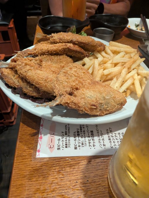 The image shows a plate of food, including fried fish and fries, placed on top of a wooden table. The plate contains several pieces of fried fish and a side of french fries. There are also two cups present on the table - one near the plate with the food and another further away from it. A person can be seen in the background, but their face is not visible due to the angle of the photo.
