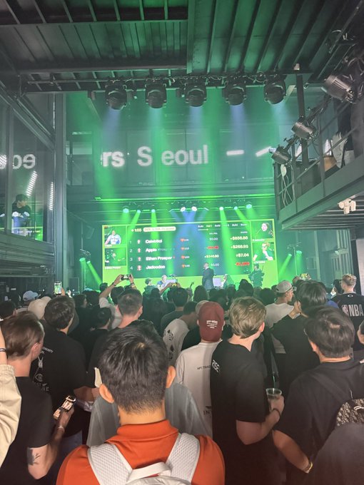 A crowd at an esports event in Seoul watches a large screen displaying player leaderboards under green stage lighting.