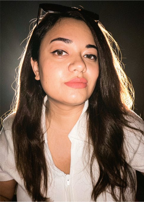A close-up selfie of a young woman with long dark hair, wearing a white zip-up top with sunglasses on her head.