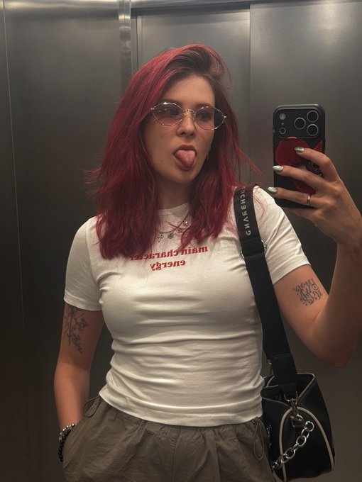 A woman with bright red hair takes a mirror selfie, sticking her tongue out while wearing a white t-shirt with reversed text and a bag strap reading 'MICHAEL KORS'.