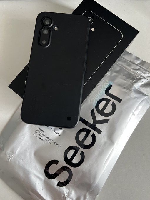A black smartphone with a triple camera setup resting on a sealed plastic bag branded 'Seeker' next to its black packaging box.