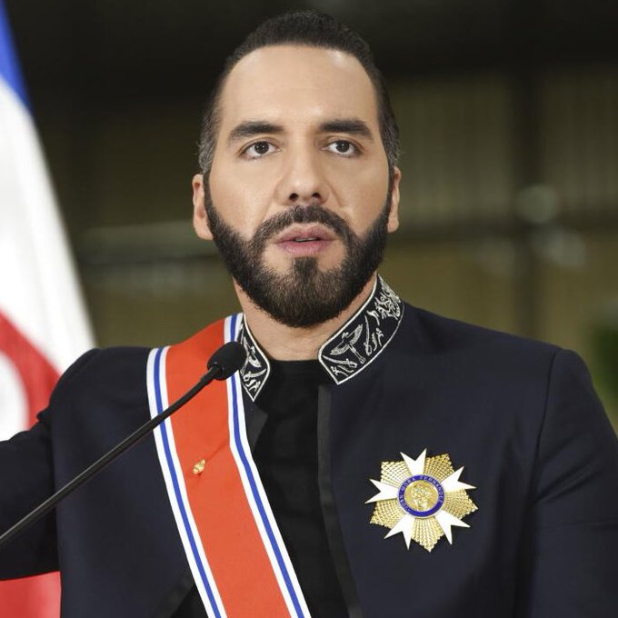 A man with a beard wearing a dark formal jacket with an ornate gold star-shaped medal and a red sash, standing before a microphone.