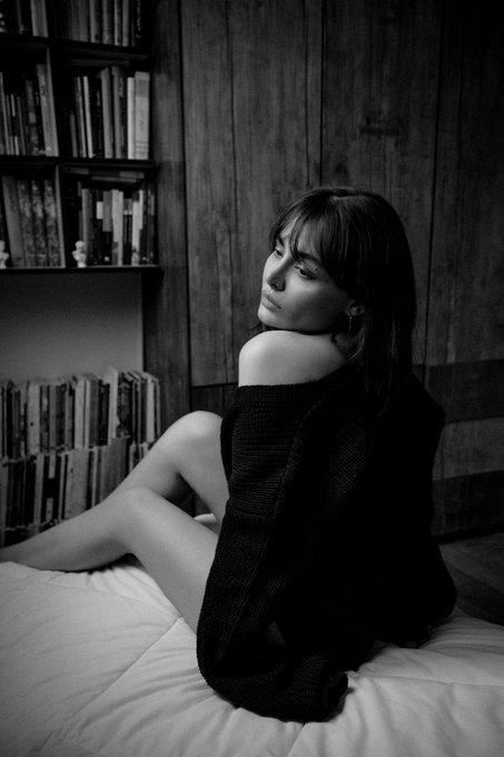 A black and white portrait of a young woman in an off-shoulder sweater sitting on a bed, looking back over her shoulder.
